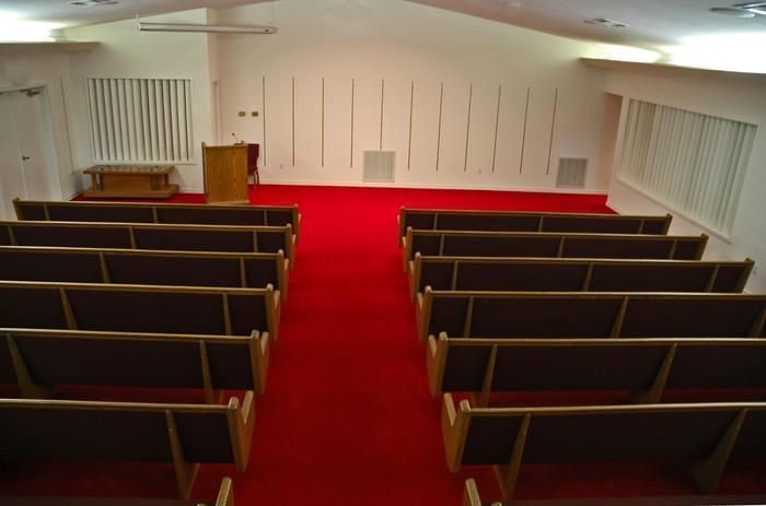 Rows of empty wooden pews face a podium and altar in a chapel with red carpet and white walls.