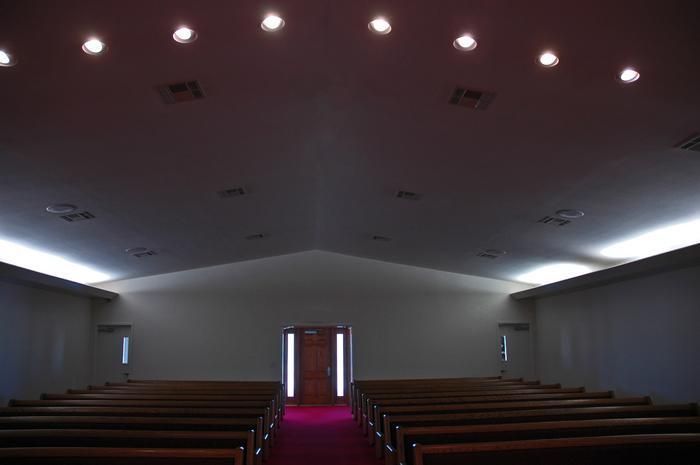 Empty church interior with rows of pews facing a doorway, red carpet, and ceiling lights.