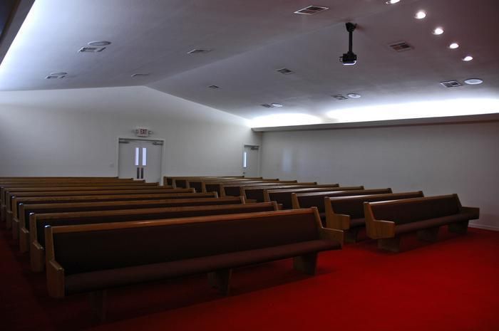Interior of a sparsely filled chapel with rows of wooden pews on red carpet.