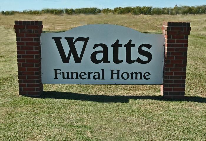 Sign for Watts Funeral Home, white with black text, flanked by brick pillars, set in a grassy field.