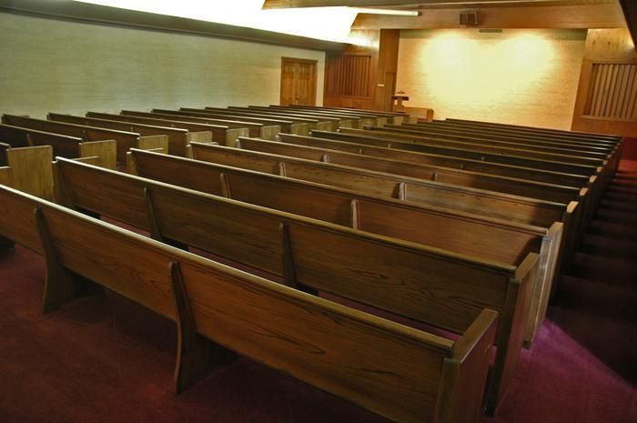 Rows of empty wooden pews in a dimly lit auditorium with a stage in the background.