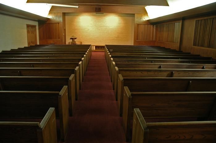 An empty church interior, rows of wooden pews facing a wooden altar, lit by warm light.