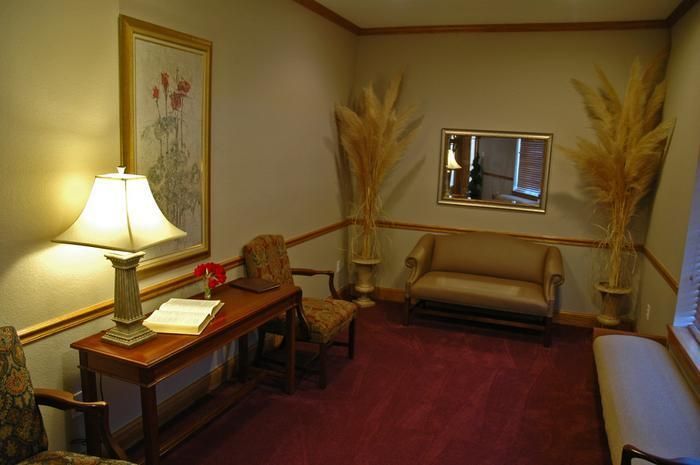 Waiting room with a loveseat, desk, mirror, and tall dried grasses. Red carpet, beige walls, and a lamp.
