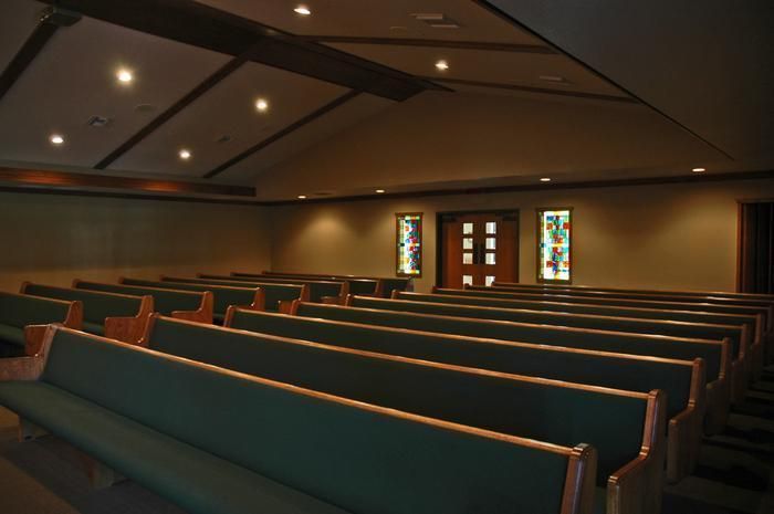 Empty chapel with rows of green-cushioned pews facing a wooden door and two stained glass windows.