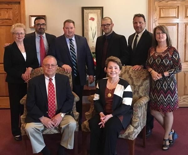 Group of nine people posing for a photo.  Two seated in chairs, the rest standing. Business attire, formal setting.
