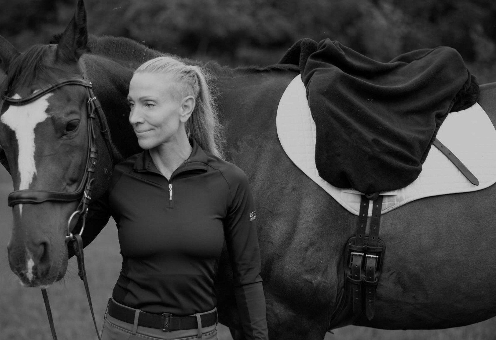 Woman in riding attire stands beside a saddled horse in a field; black and white.