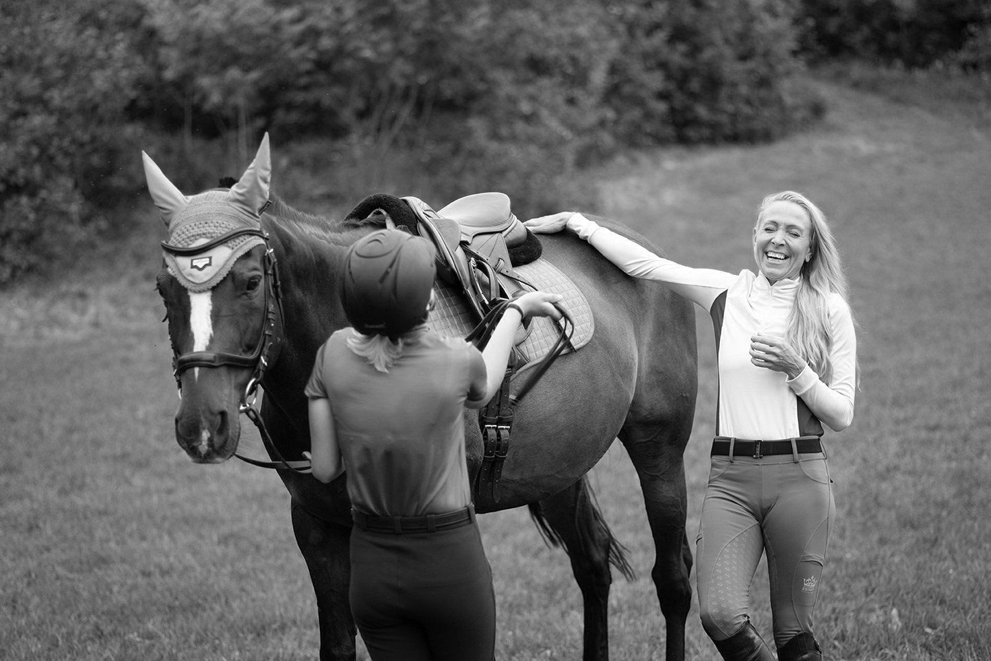 Two women and a saddled horse in a field. One woman adjusts the saddle, the other laughs and pets the horse.