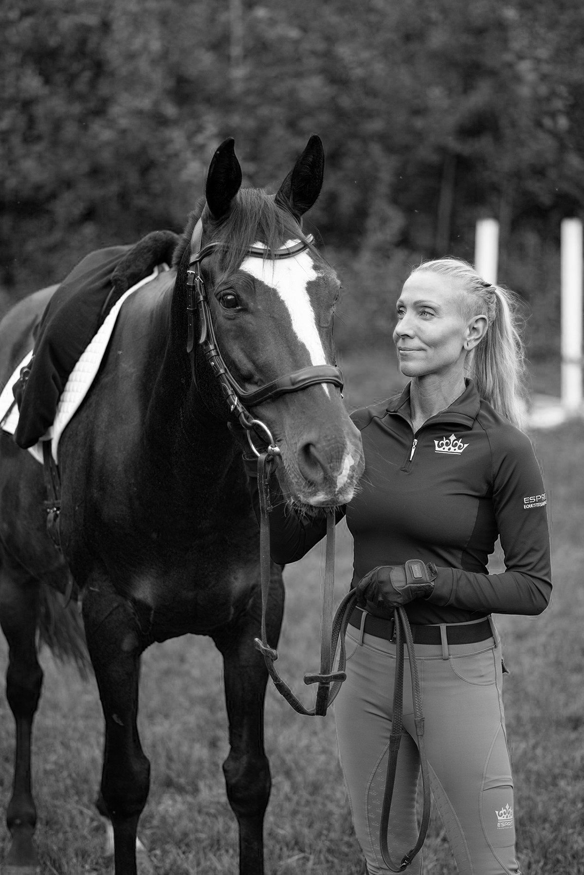 Woman with blonde hair smiles, holding reins of a dark horse with a white blaze in a field.