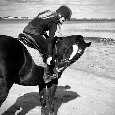 Woman on horseback on a beach, wearing helmet. Horse is dark, on sand, near water.