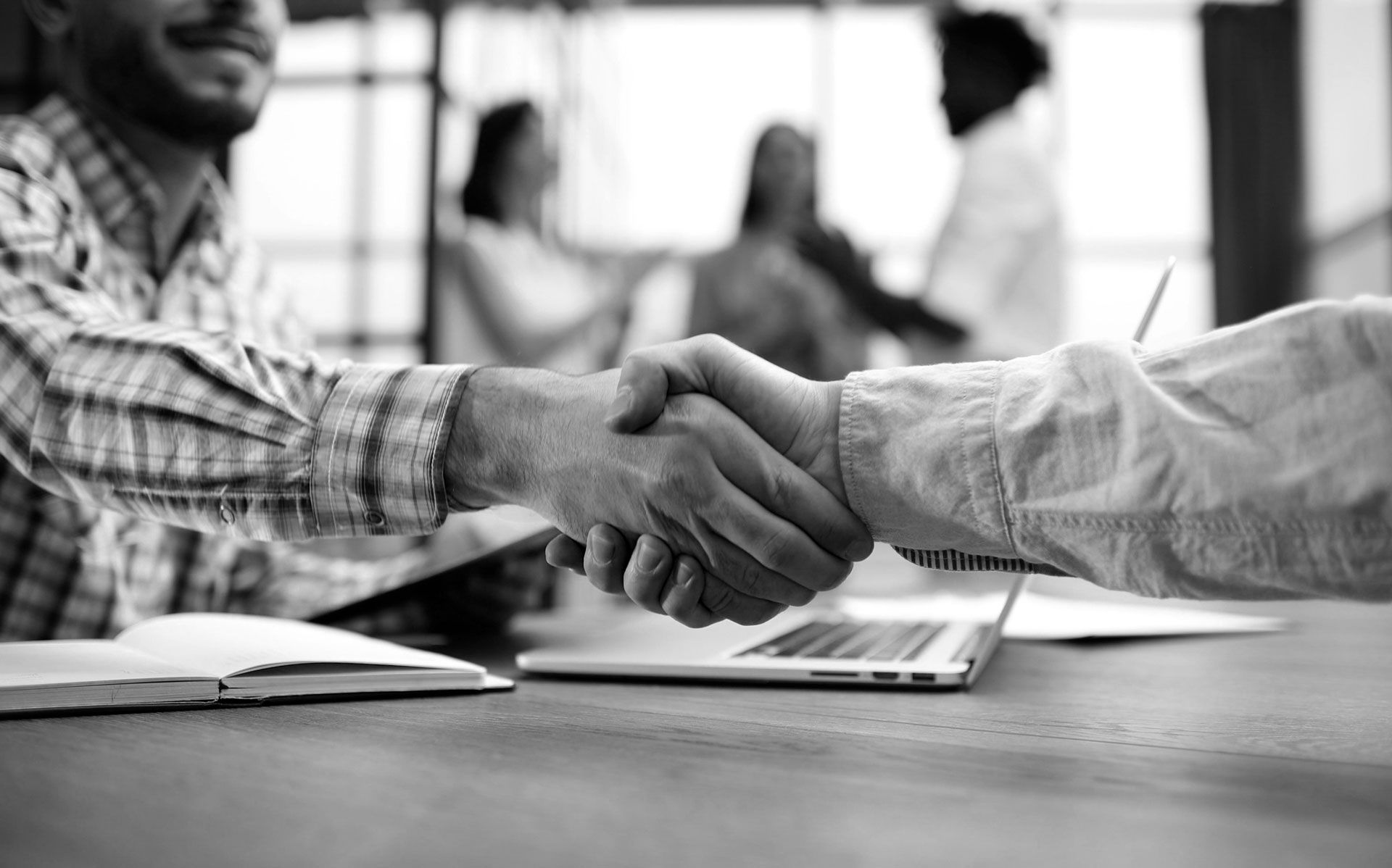 Two people shaking hands at a table, others blurred in the background, business meeting.