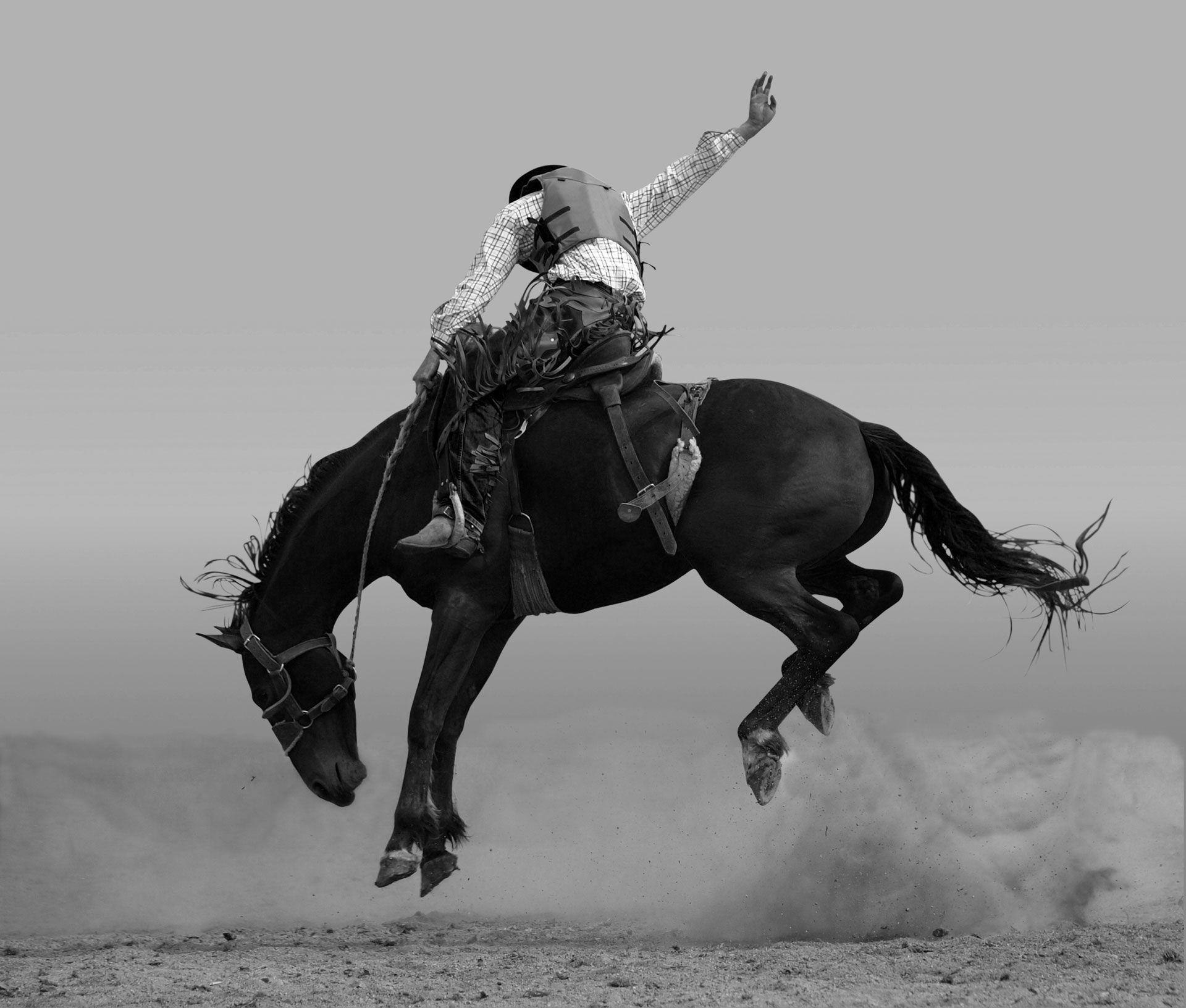 Rodeo rider on bucking horse, dust in the air; rider in mid-air with one arm raised.