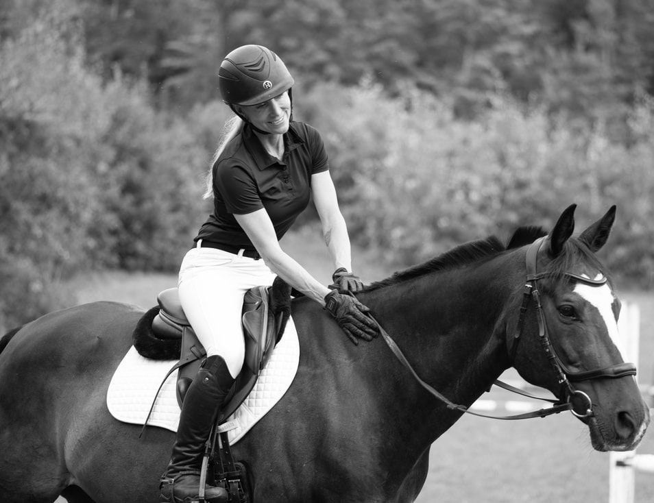 Woman on horseback, wearing riding helmet and gloves, petting the horse's neck.