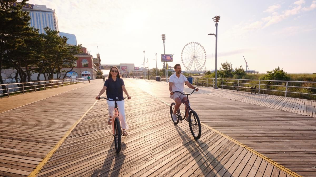 Couple riding bikes on a boardwalk; Ferris wheel and buildings in the distance.