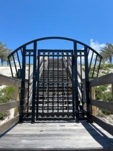 Black gate with arched top blocks stairs to beach. Wooden ramp leads to the gate. Blue sky.