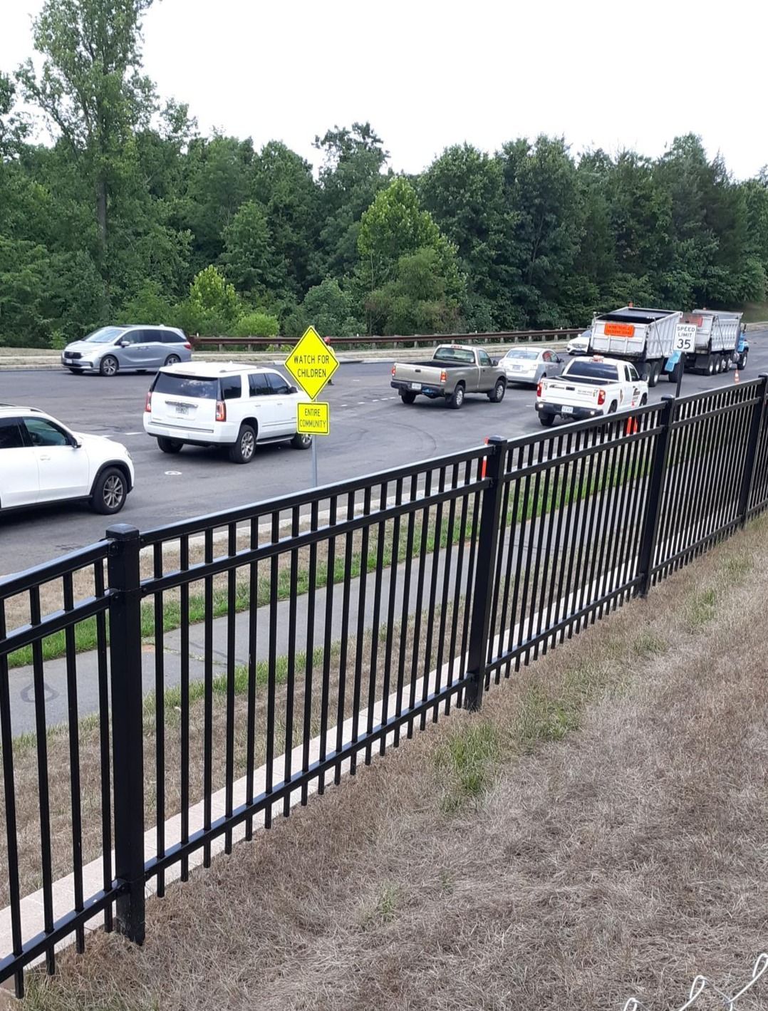 Black fence in foreground with traffic in the background. Cars and trucks on the road, surrounded by trees and greenery.