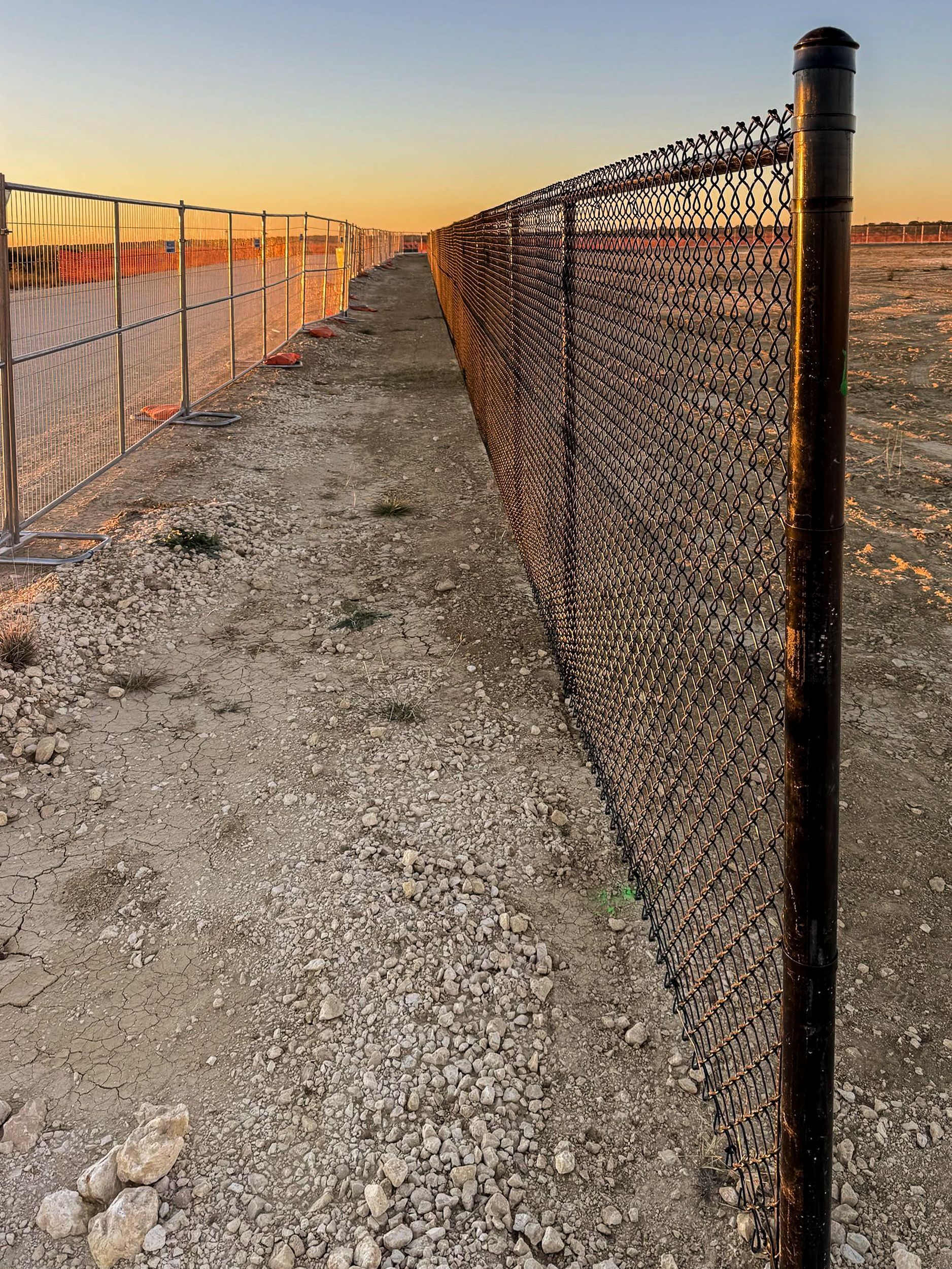 Gravel path between two fences under a sunset sky. One fence is metal and one is temporary construction fencing.