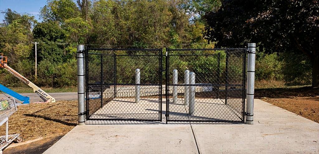 A fenced-in utility area with gray concrete, pipes, and green trees in the background.