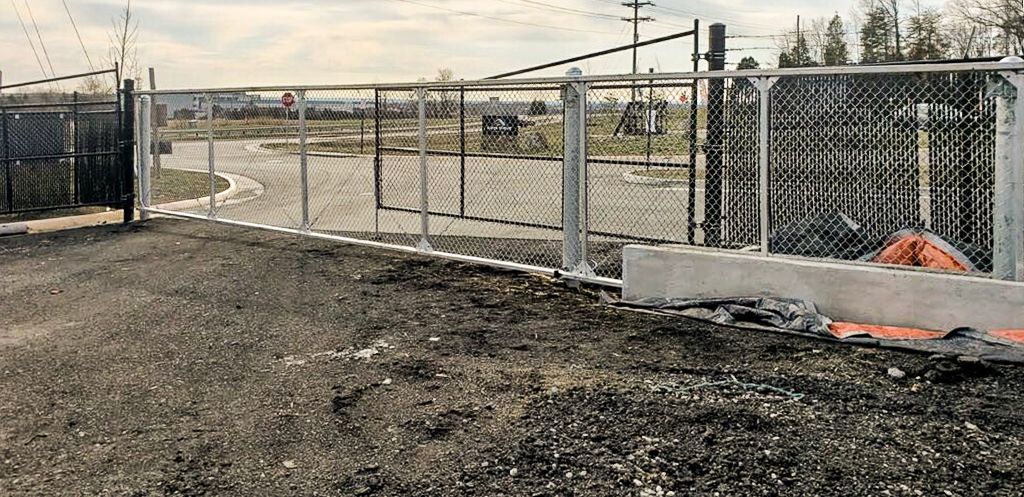 A chain-link fence surrounds a gravel area; a gate is open, and a building is in the distance.