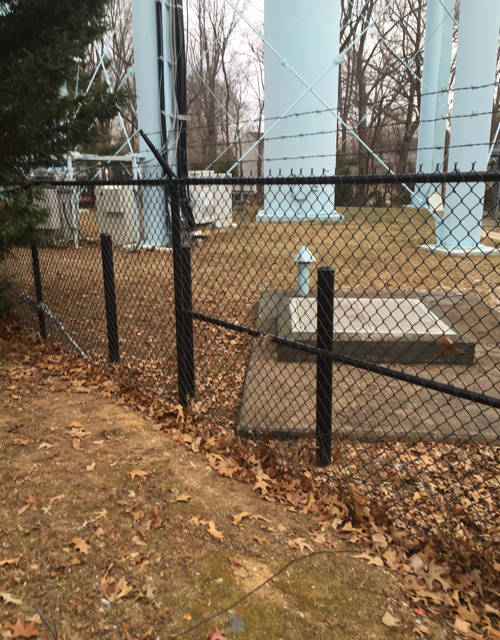 Black chain-link fence surrounds the base of water towers in an outdoor setting. Brown leaves and grass present.