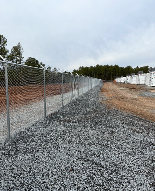 Chain-link fence with barbed wire atop, bordering a gravel path and dirt area under a cloudy sky.