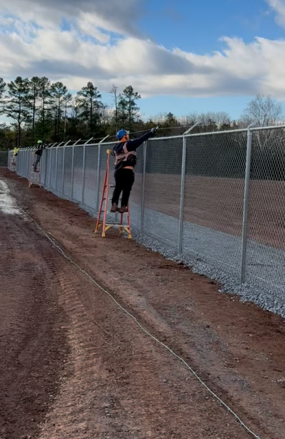 Two people installing security fence with barbed wire on a ladder, outdoors.