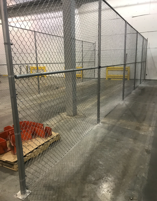 Chain-link fence enclosure within a warehouse. Concrete floor, metal supports, and orange objects.