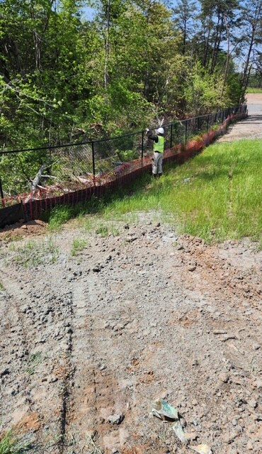 A person in a safety vest stands near a new black fence built on a brick wall alongside a road; trees in the background.