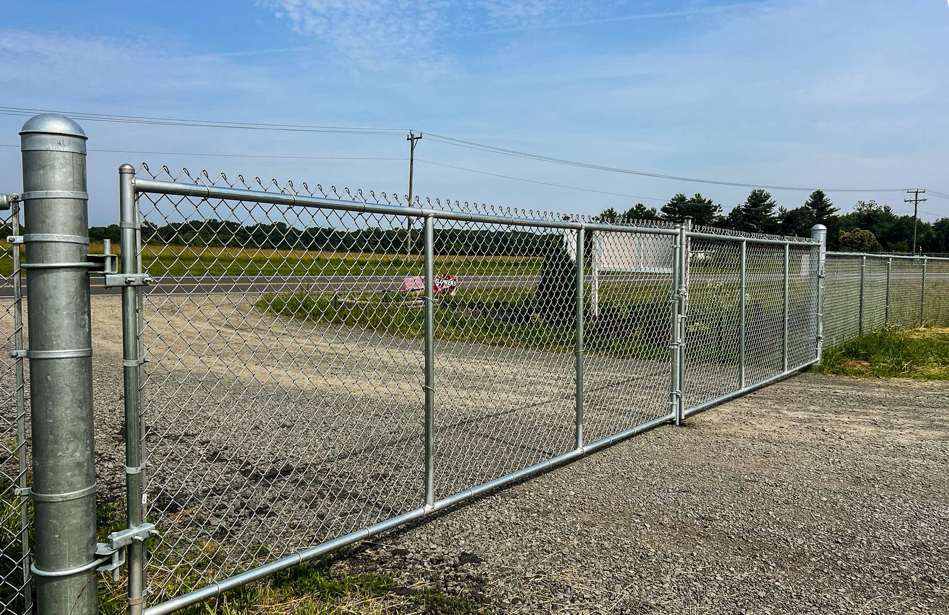 Chain-link fence with a gate, in a gravel parking lot, under a blue sky.