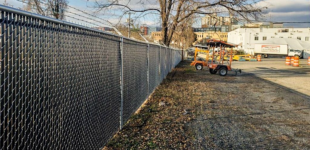 Chain-link fence with barbed wire atop. A construction site beyond the fence with orange vehicles.