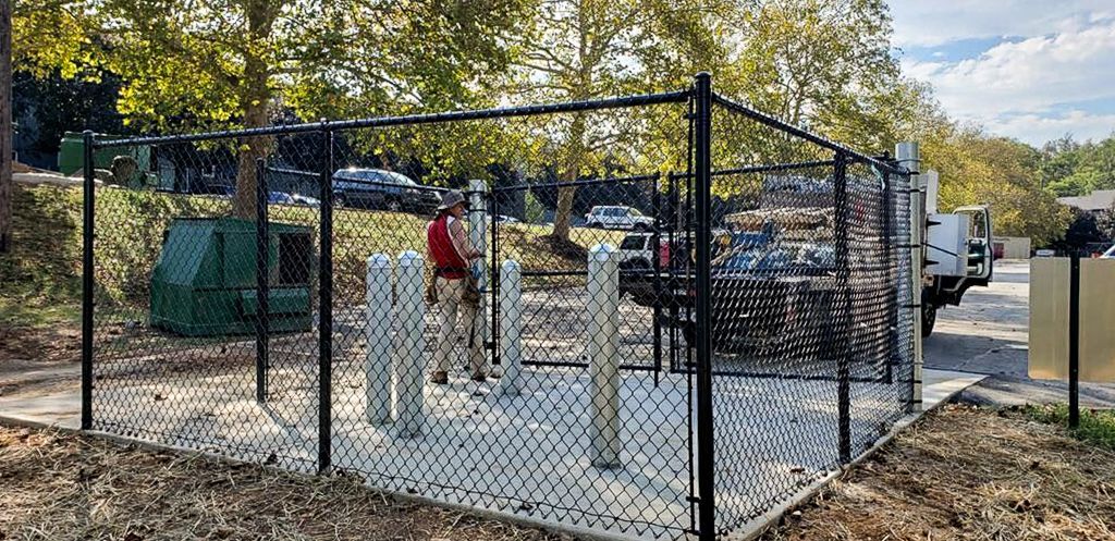 A chain-link fence surrounds concrete pillars. A person stands inside the fenced area near the pillars. Trees and a truck are in the background.