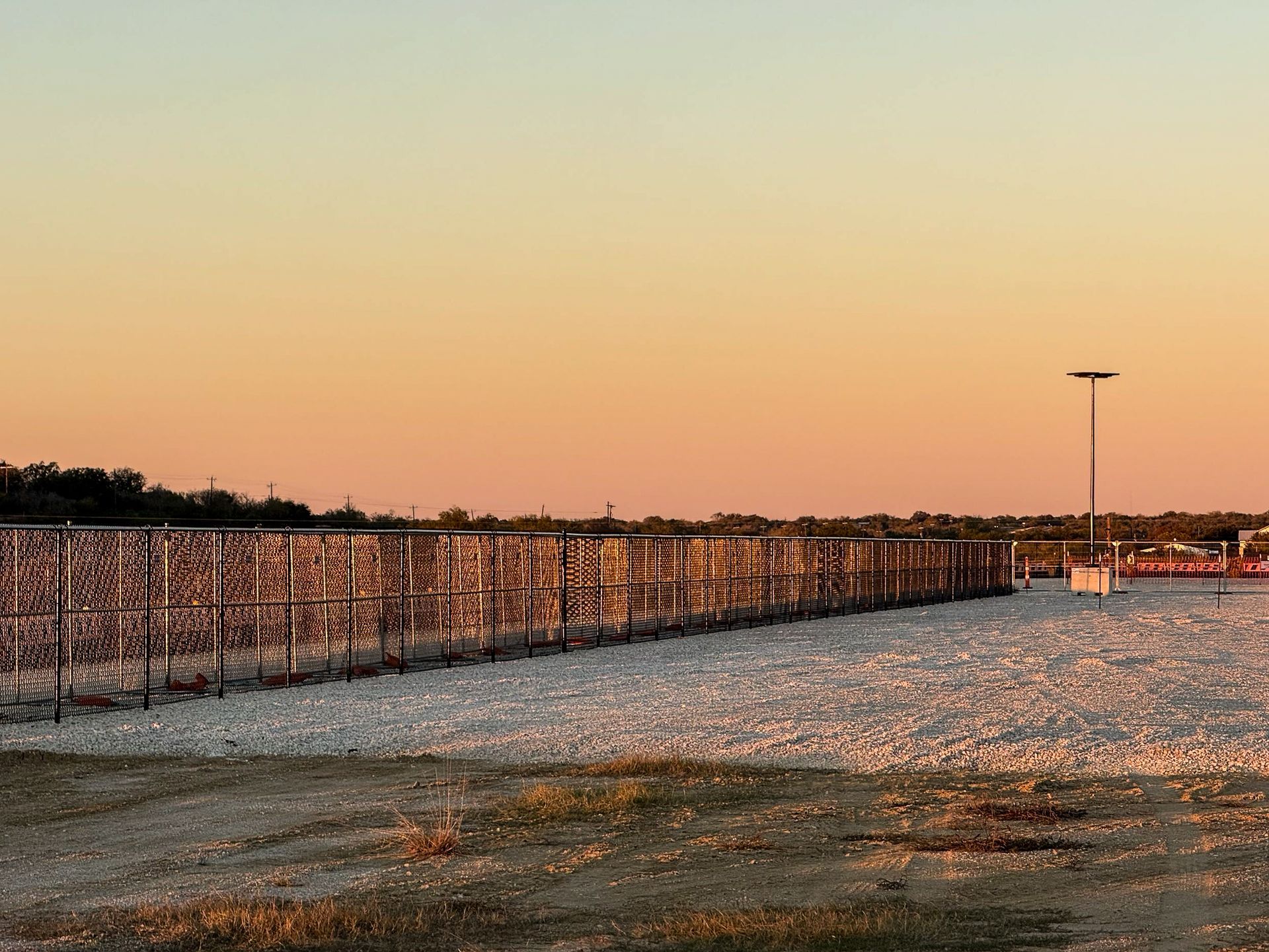 A long fence made of a trellis is on a rocky, beige ground at sunset with a street light in the distance.
