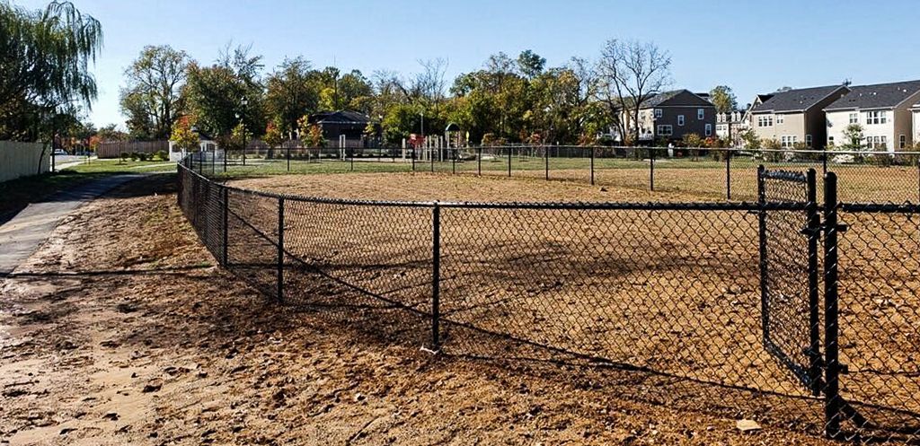 Black chain-link fence encloses a dirt area, trees in the background, houses beyond. Sunny day.