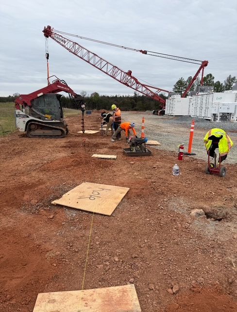 Construction site with crane, workers, and machinery on reddish-brown ground; overcast sky.