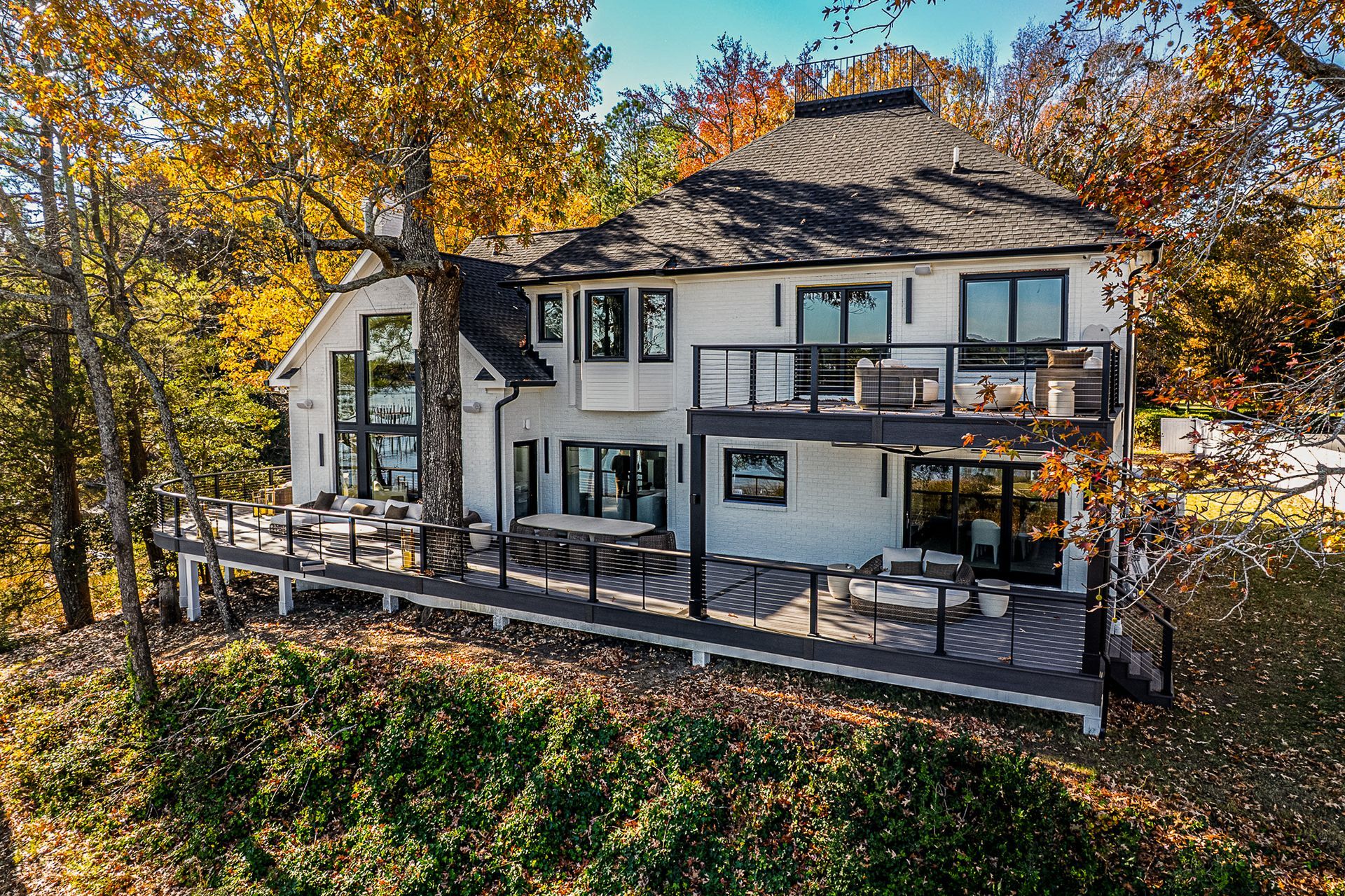 White house with deck overlooking a lake, surrounded by autumn trees.