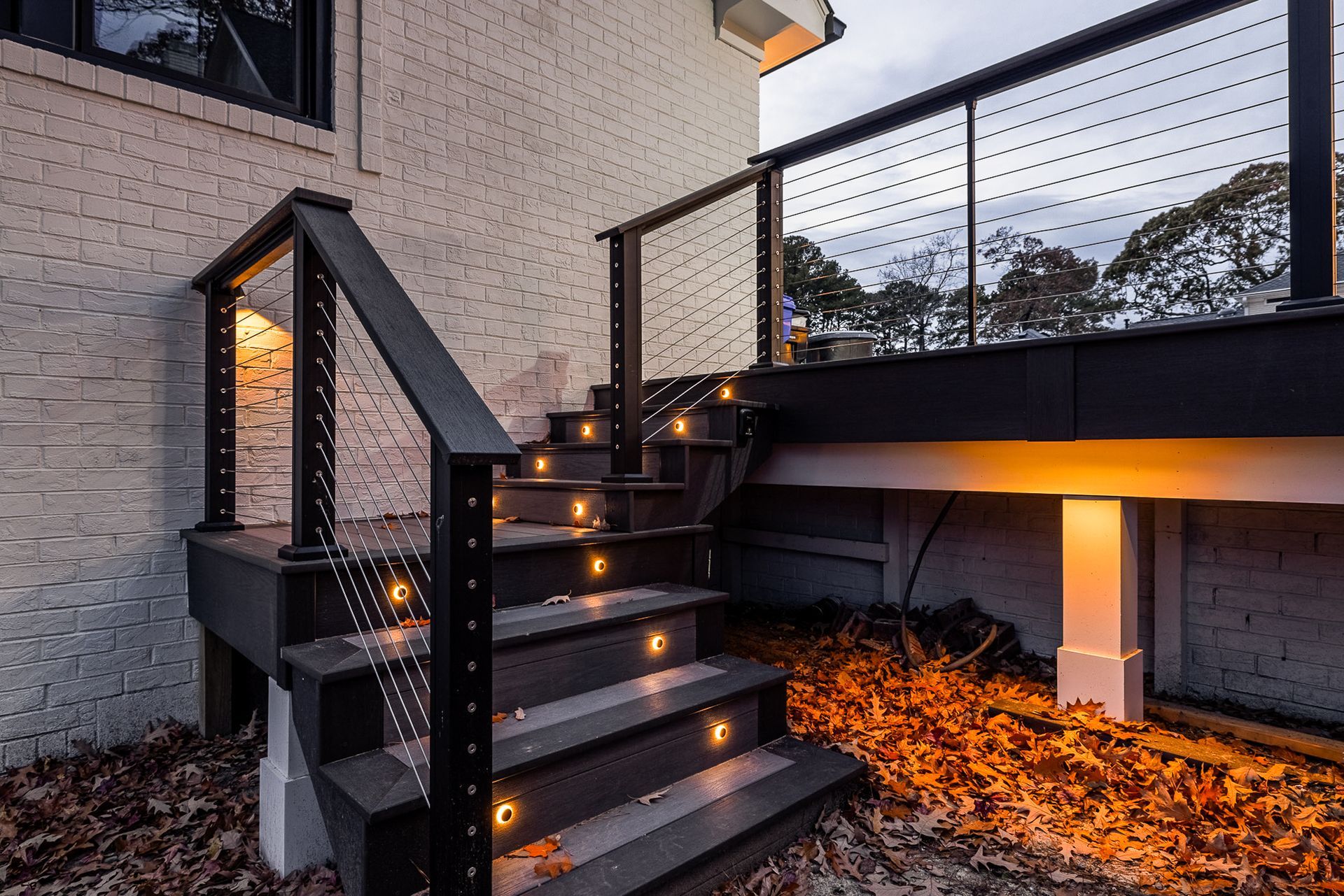 Dark gray outdoor stairs with built-in lights, railing, and leading to a deck near a brick building.