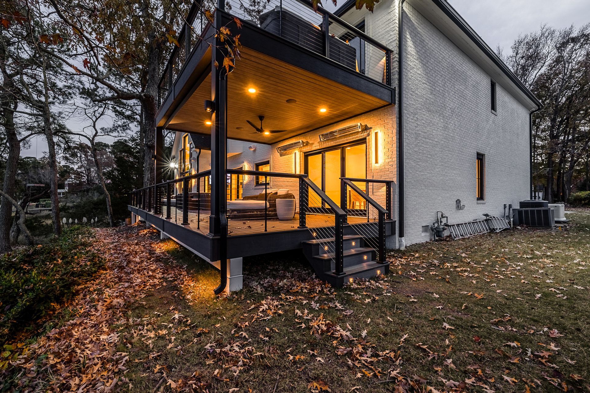 Two-story home with lit deck and stairs, surrounded by fall foliage.