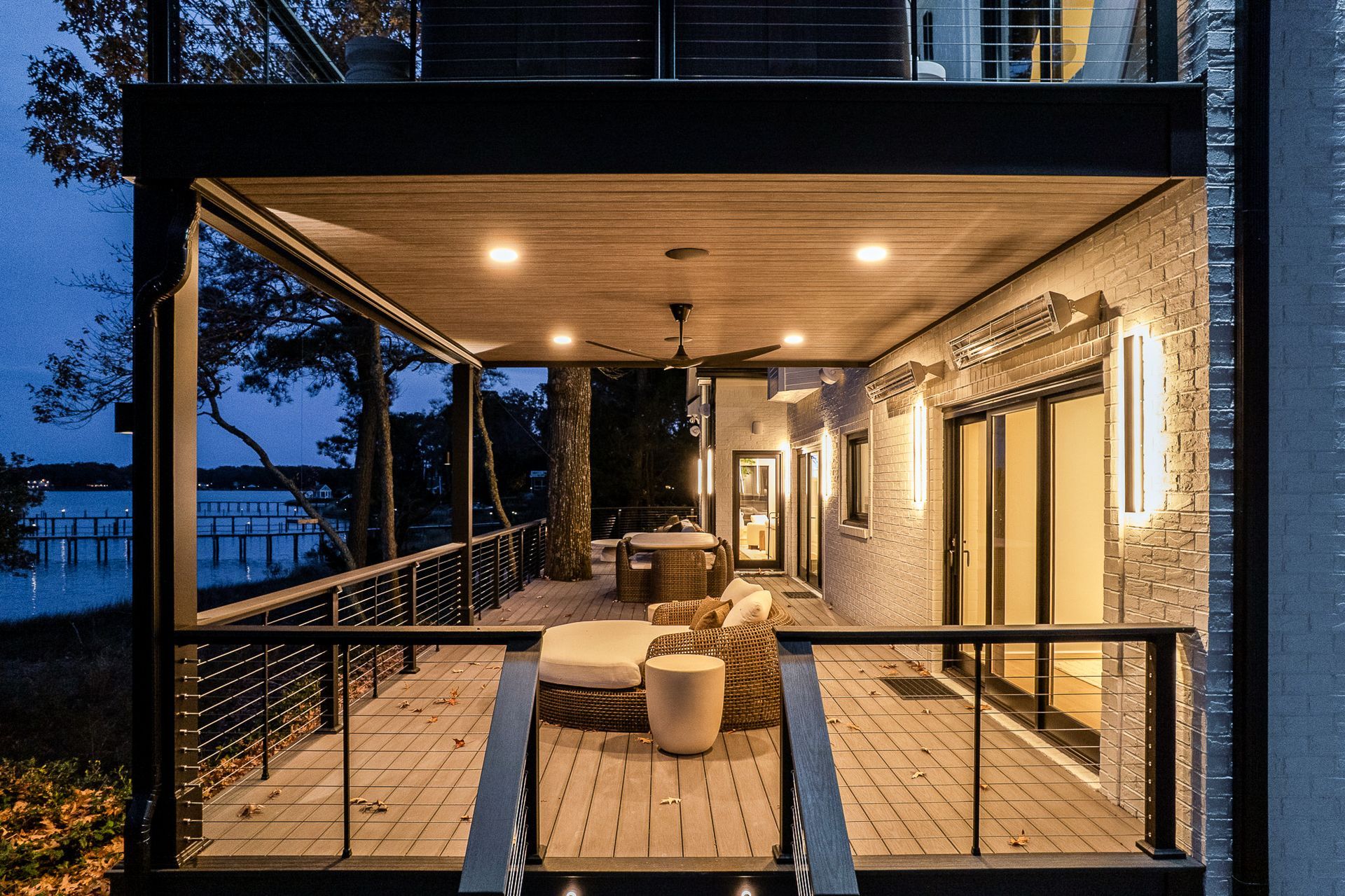 Outdoor deck with seating, overlooking water at dusk. Lit by recessed lights and sconces.