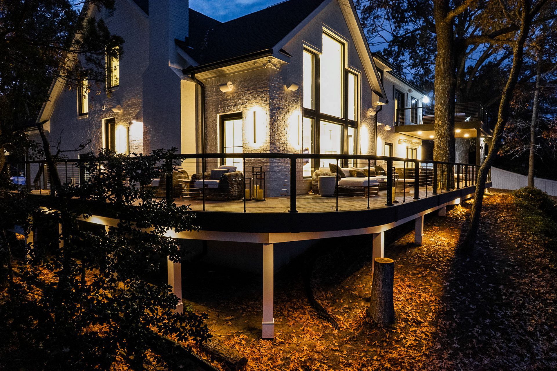 Lit-up modern home with a deck at dusk. The white house has large windows, and warm lights illuminate the deck and surrounding trees.