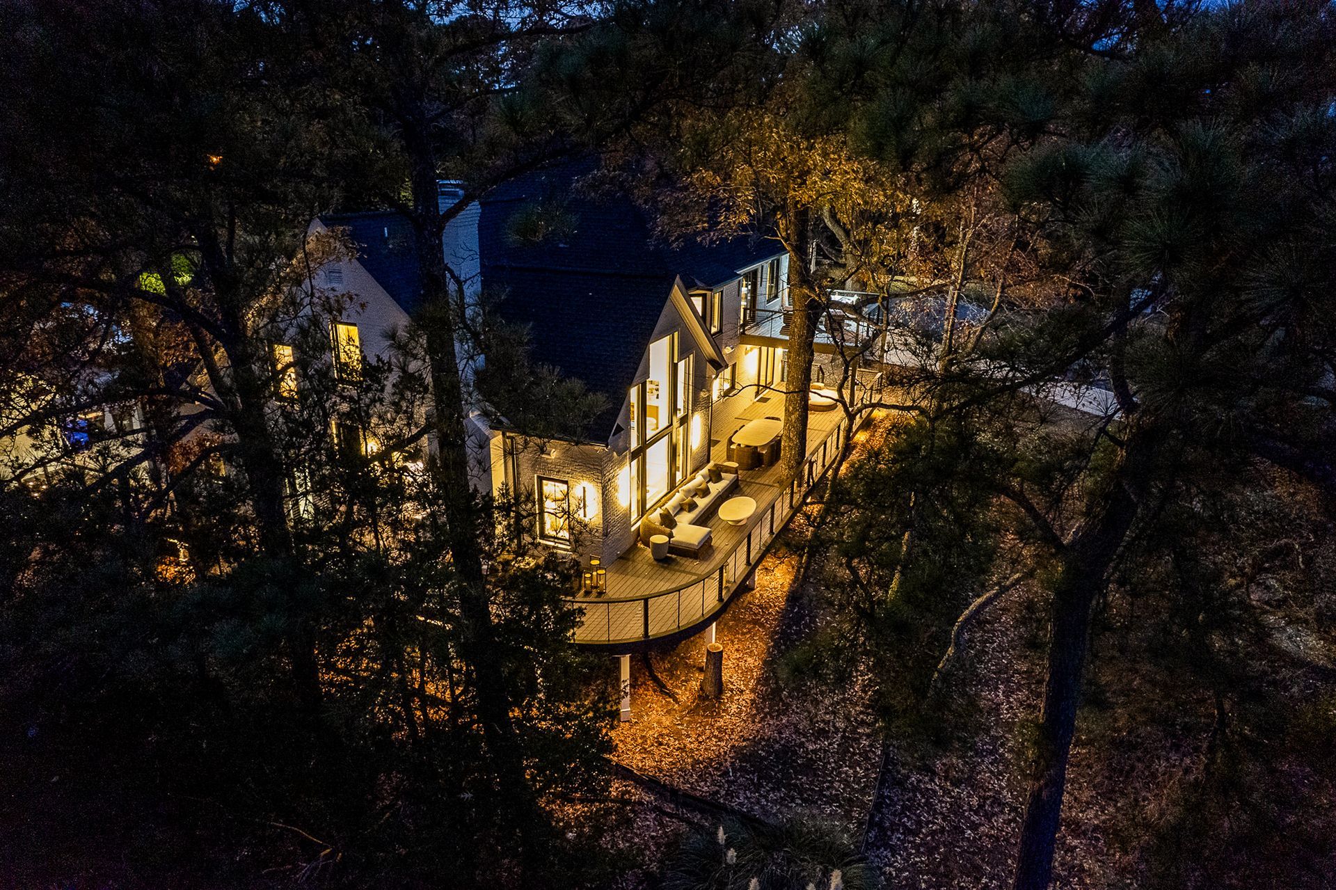 Night view of a house with lit windows and a deck, surrounded by trees.