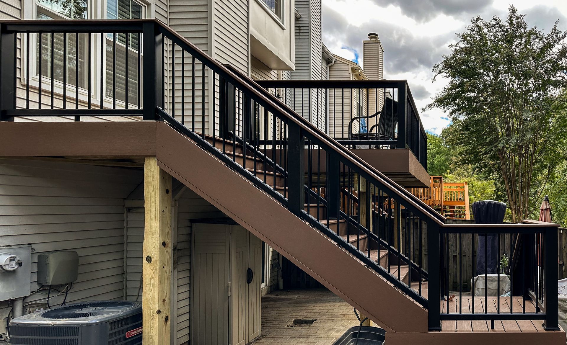 Two-level deck with black railing and brown boards, stairs leading down. Air conditioning unit visible. Cloudy sky.