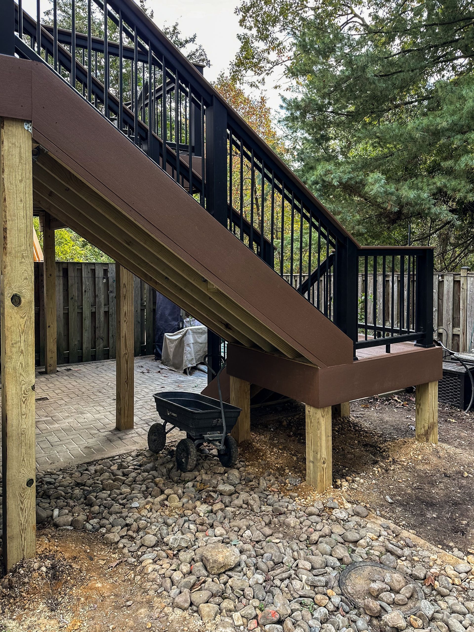 Brown wooden deck stairs with black railings, supported by posts, over gravel. A small grill sits underneath.