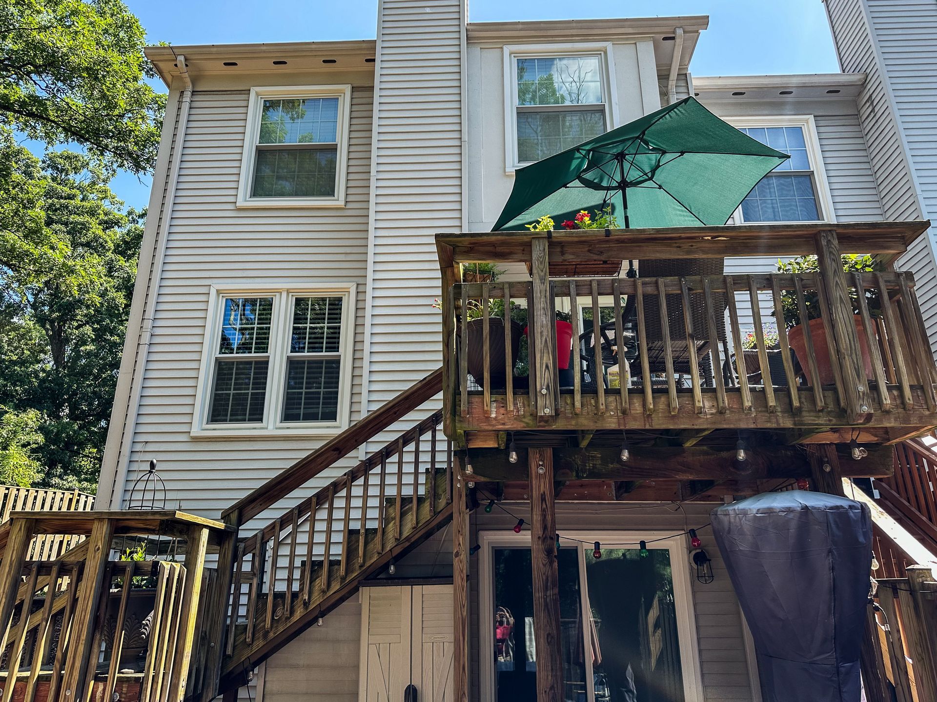 Back of a multi-story house with a wooden deck and stairs, green umbrella, and window.