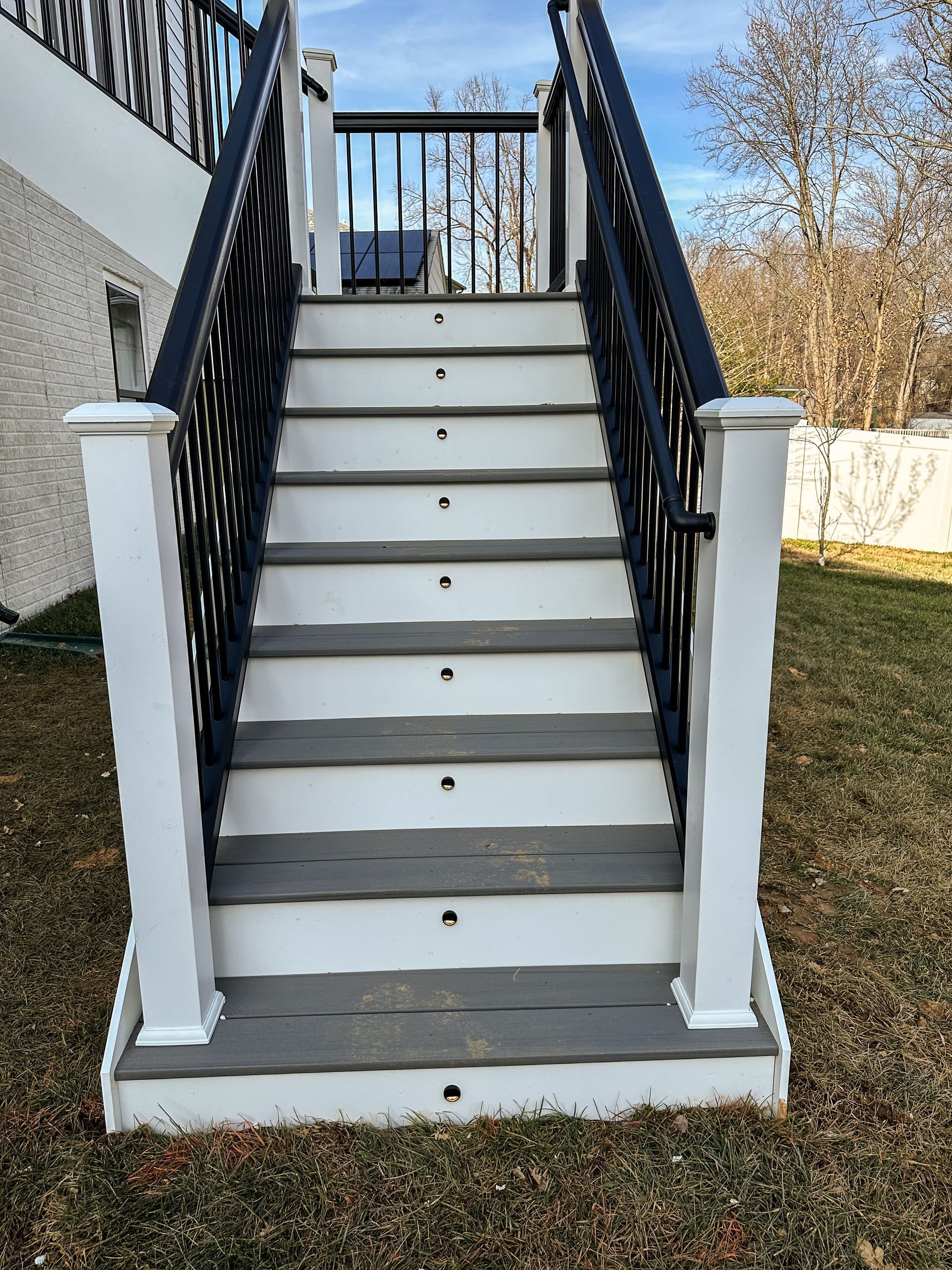 Staircase leading up to a deck with white posts, gray steps, and black railing. Set outdoors in a yard.