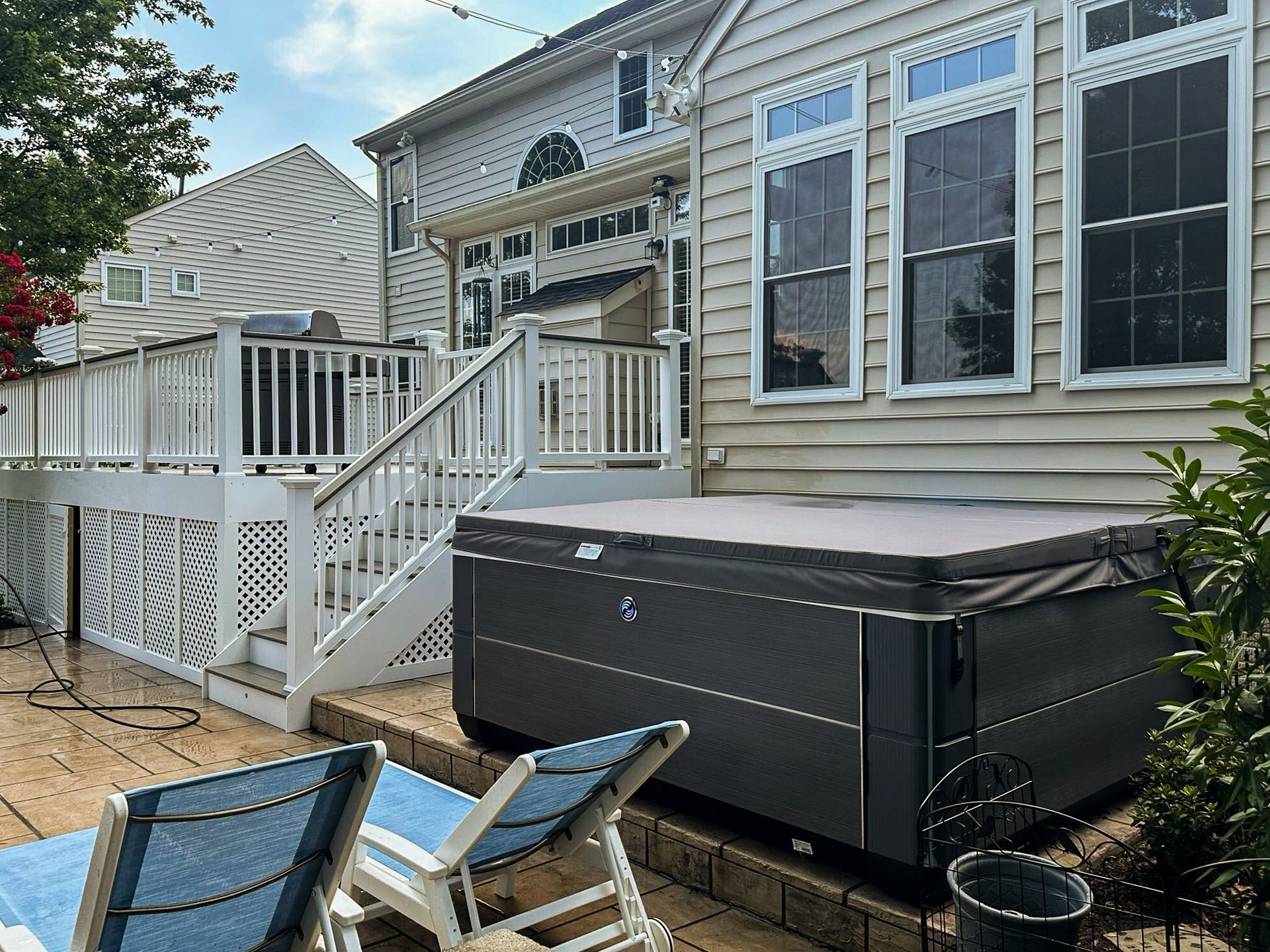 Backyard with hot tub on a patio next to a white deck, blue lounge chairs in front.