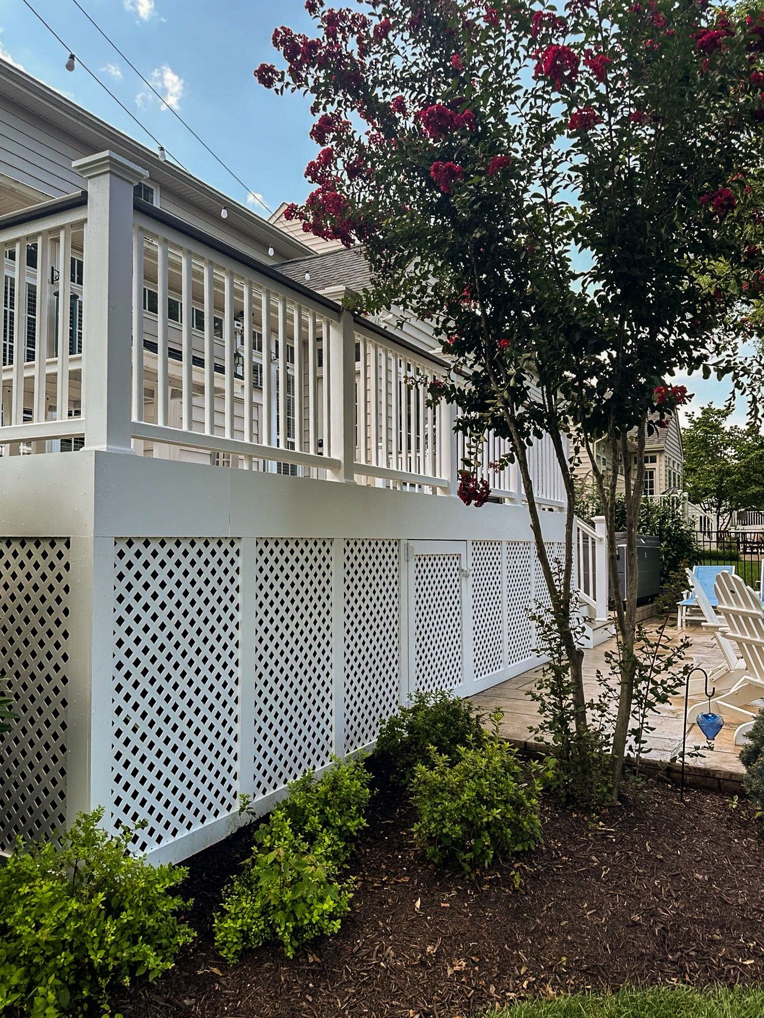 White deck with lattice skirting, small bushes, and a tree with red flowers.