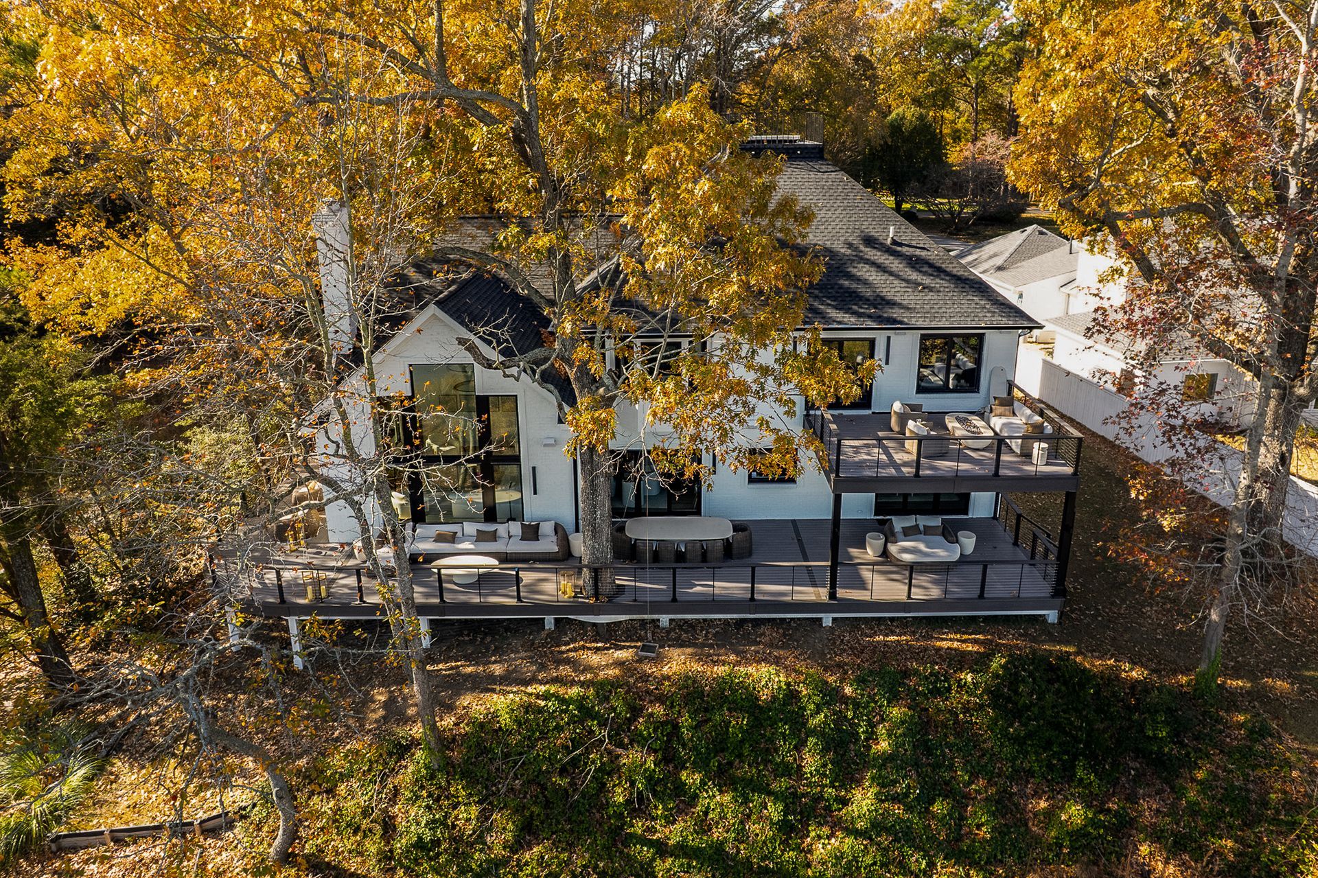 White house with black roof, multiple decks, surrounded by trees with yellow leaves.