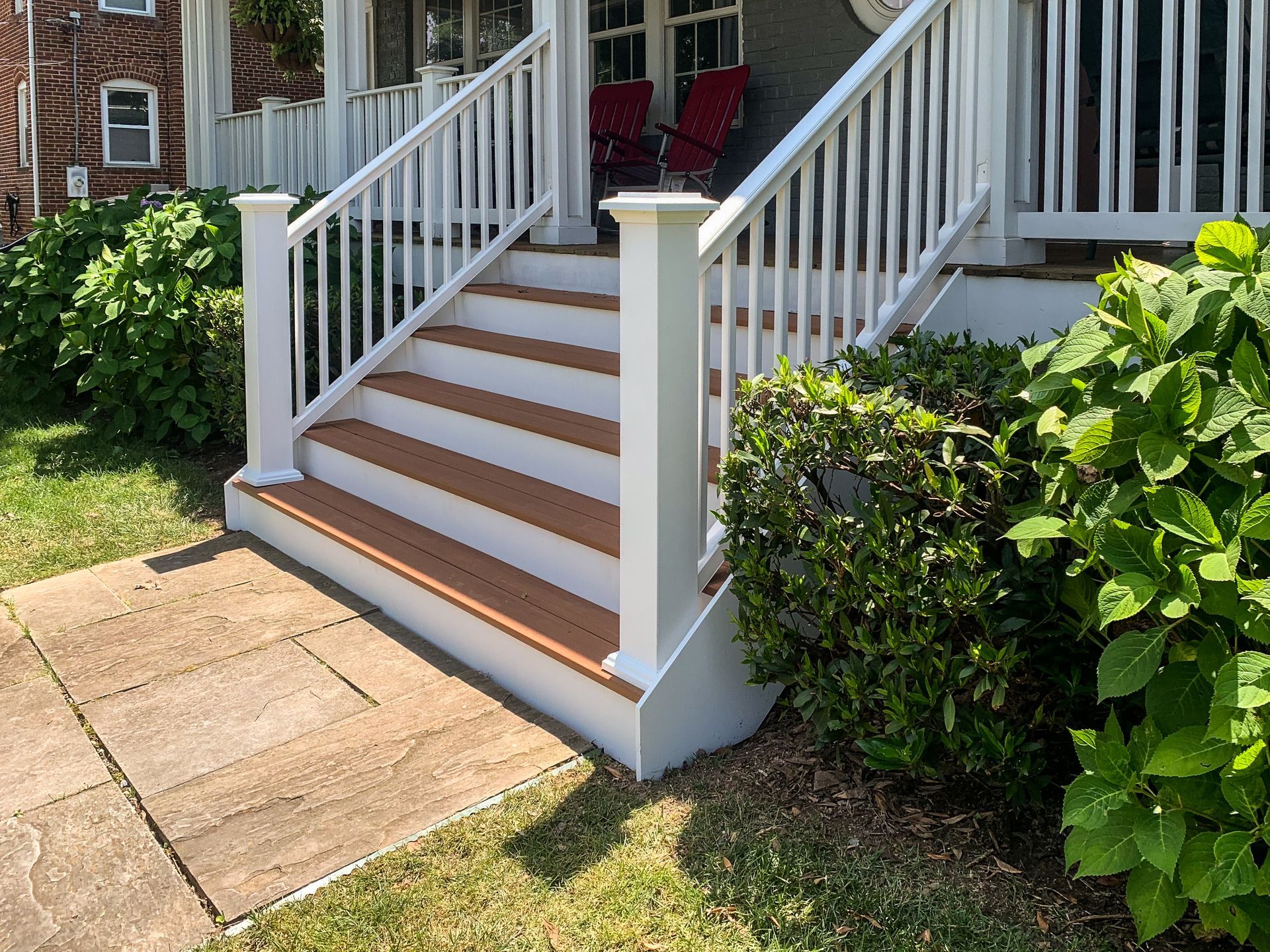 White-railed stairs with brown treads lead up to a porch. Bushes flank the stairs, fronting a brick house.