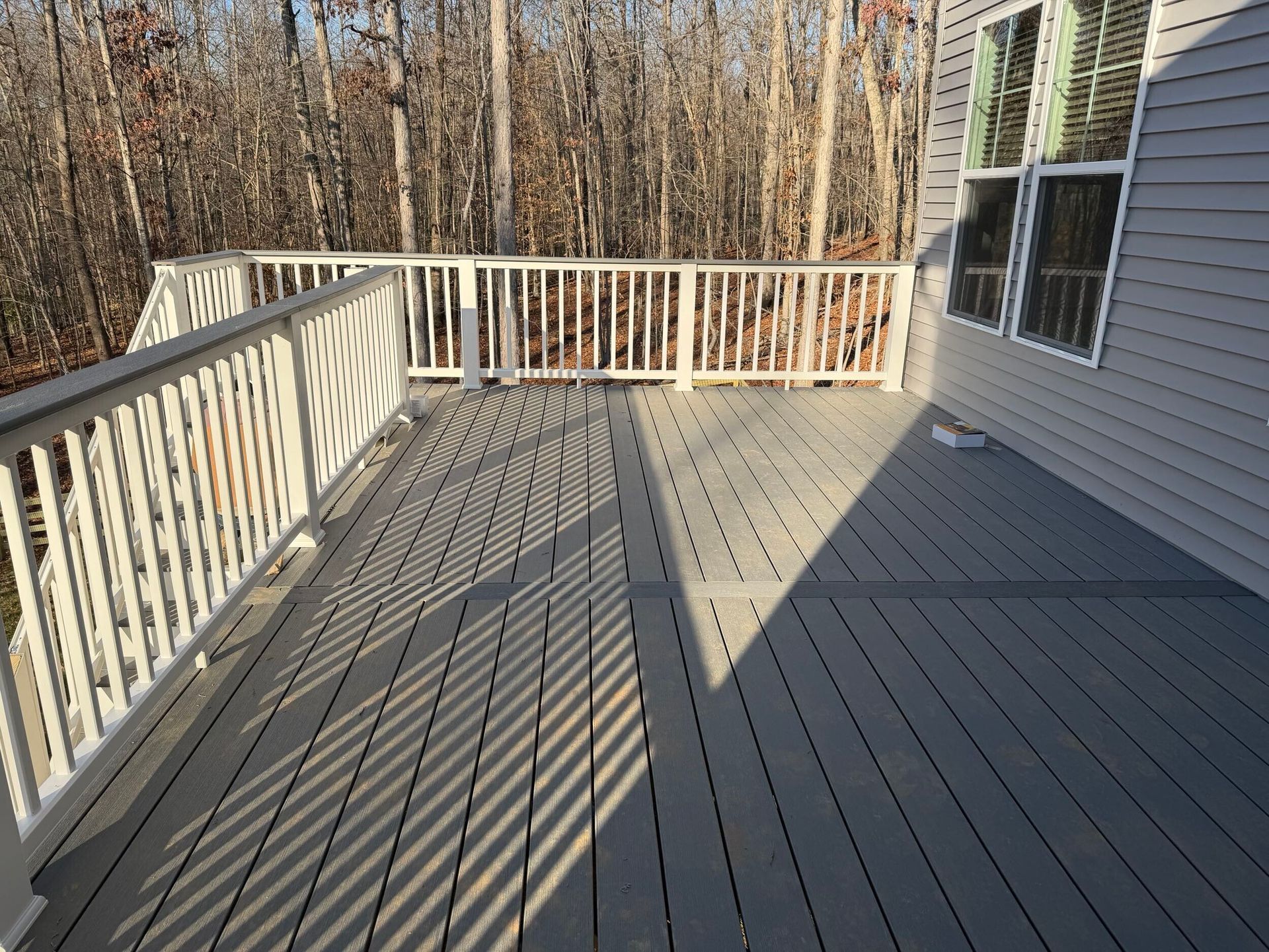 Deck with gray composite decking and white railing, attached to a house with gray siding. Sunlight casts shadows.