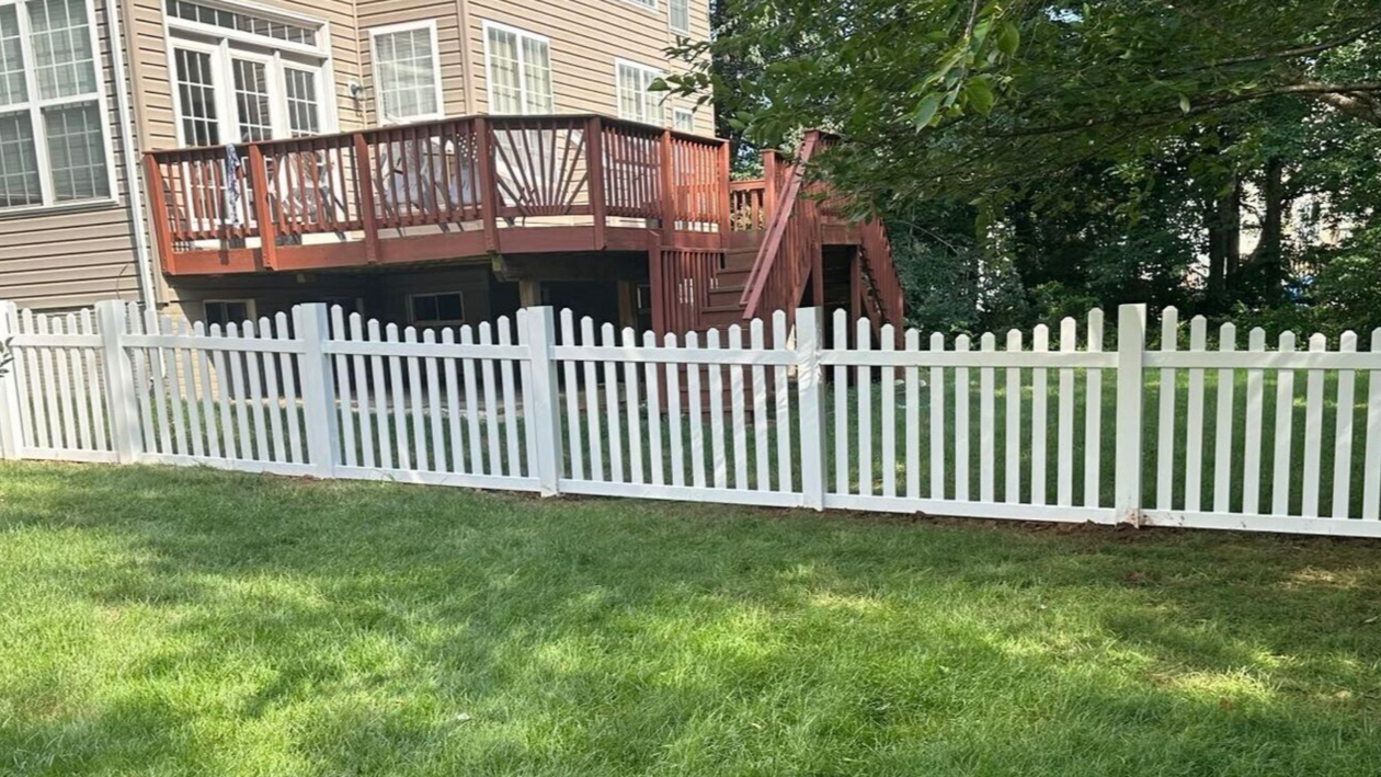 White picket fence surrounds a green lawn in front of a raised wooden deck attached to a brick building.