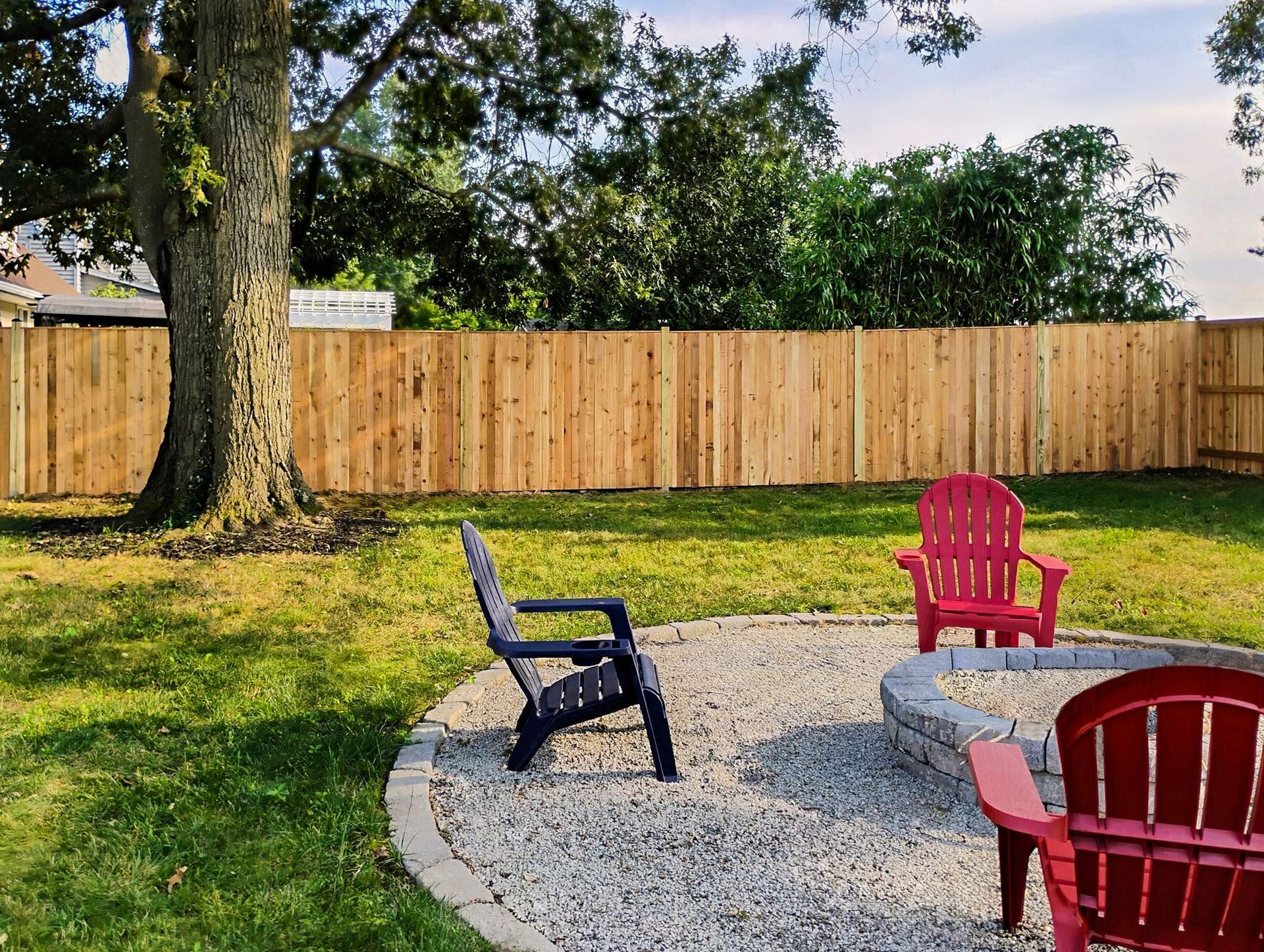 Backyard scene with gravel fire pit, red and black Adirondack chairs, wood fence, and large tree.